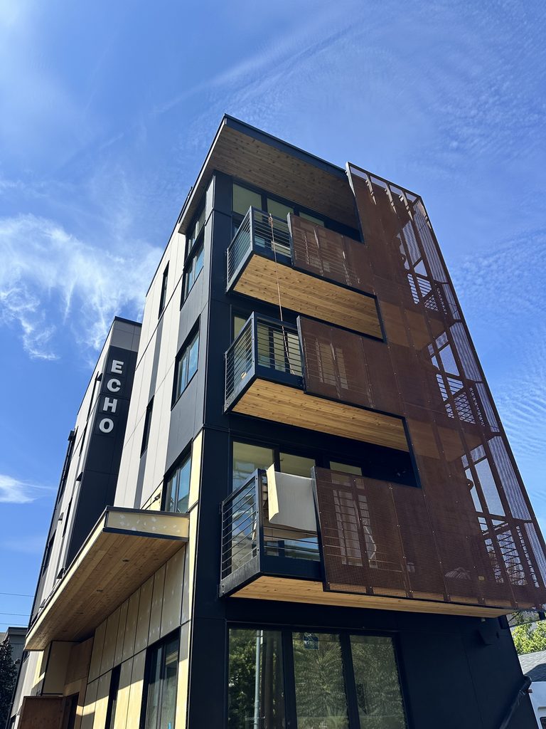Low-angle exterior view of the ECHO apartment building in Seattle, featuring dark paneling, natural wood balcony ceilings, and wire mesh railings.