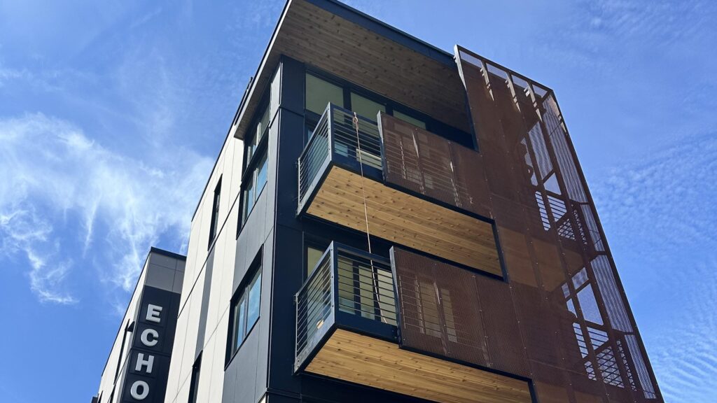 Low-angle exterior view of the ECHO apartment building in Seattle, featuring dark paneling, natural wood balcony ceilings, and wire mesh railings.