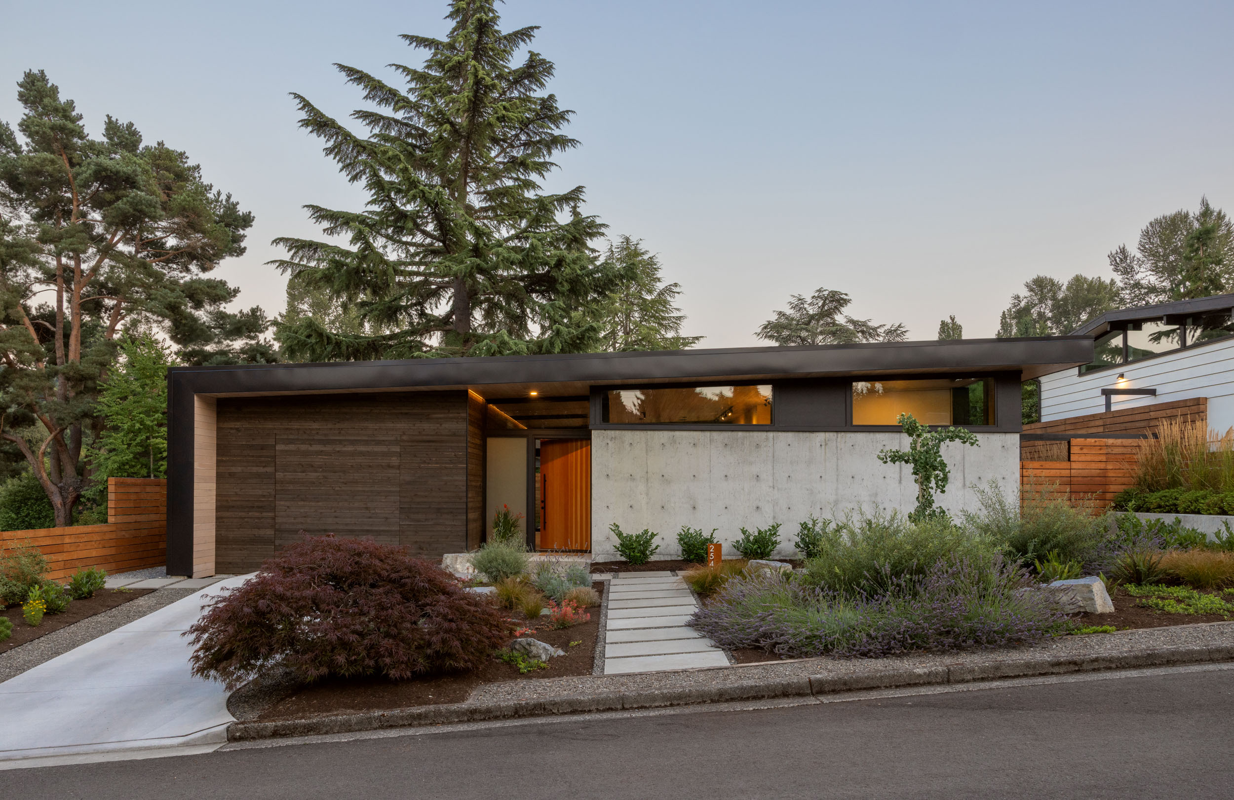 Dusk view of a modern house with wood, glass, and concrete with clean landscaping and lighting.
